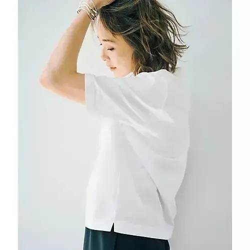 白いシャツを着た女性が横を向いている写真。茶色の短髪で、右手を頭に添えている。背景は白。黒いパンツを着用。